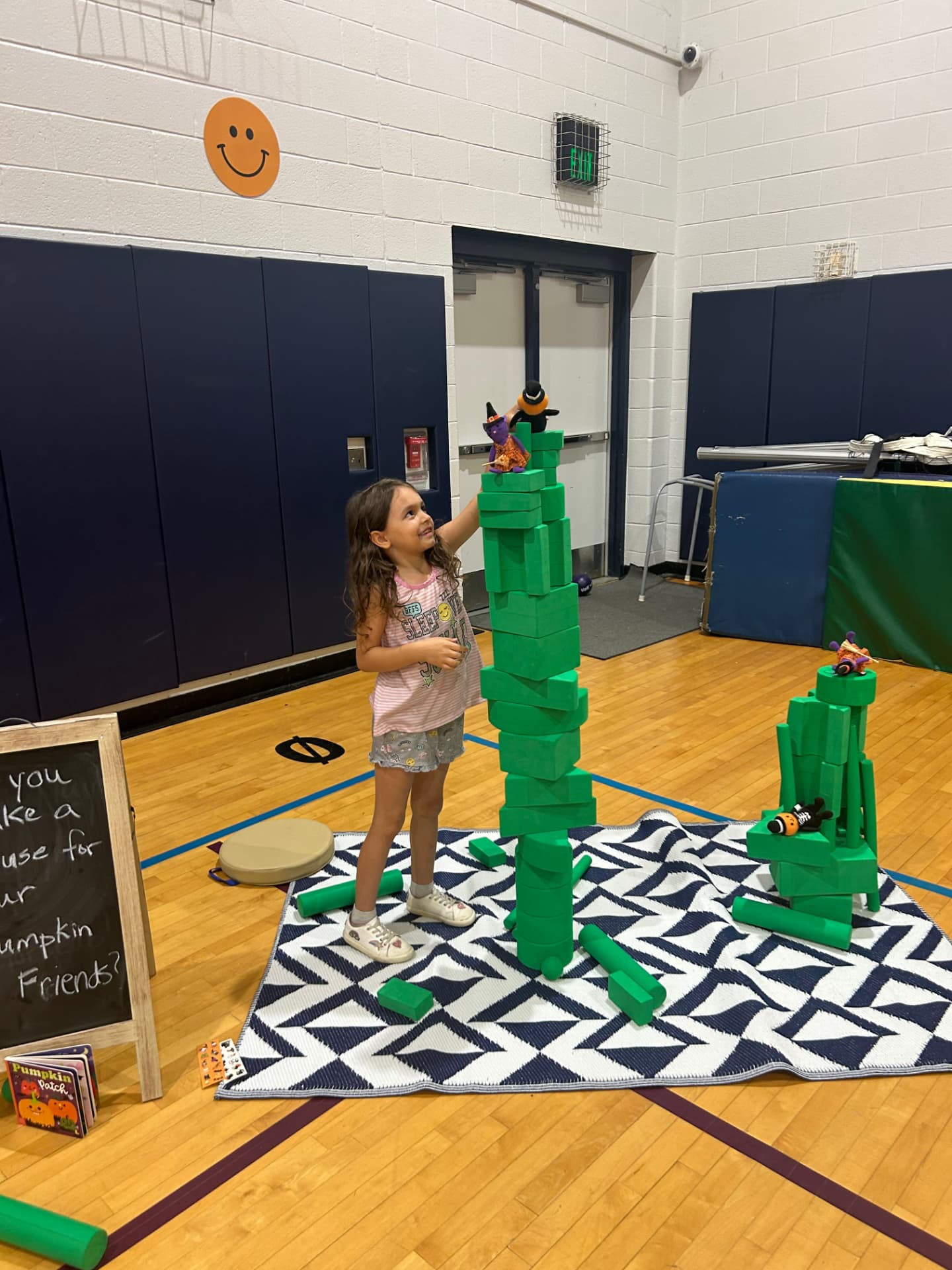 A young girl standing beside a tall tower she built from green foam blocks, with small pumpkin characters perched on top