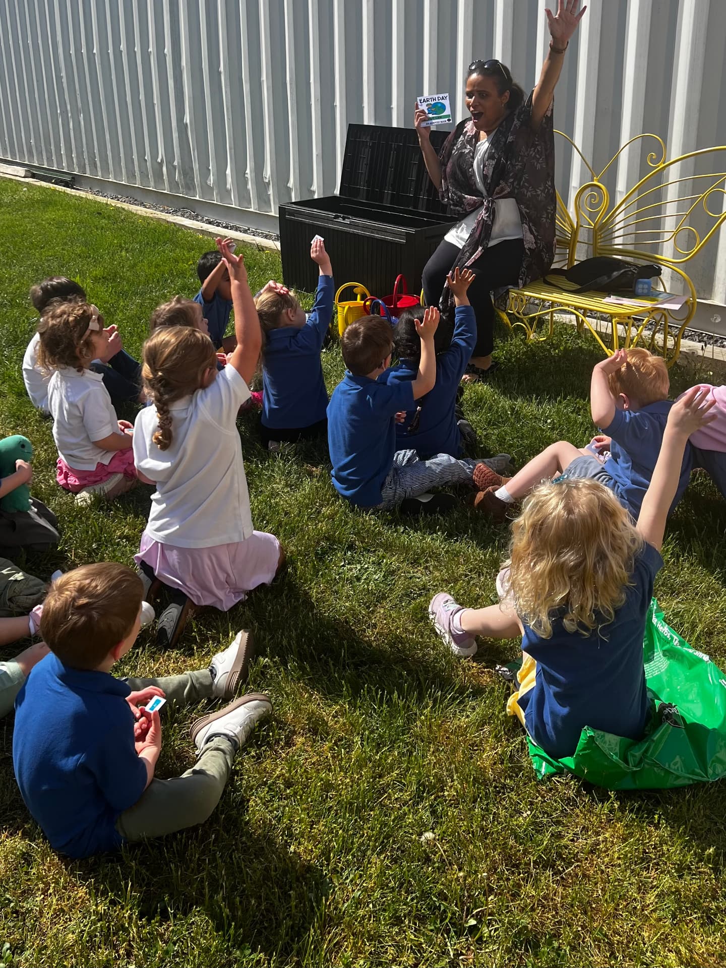 A WonderPlay host reading a story to a group of children raising their hands outdoors on the grass.